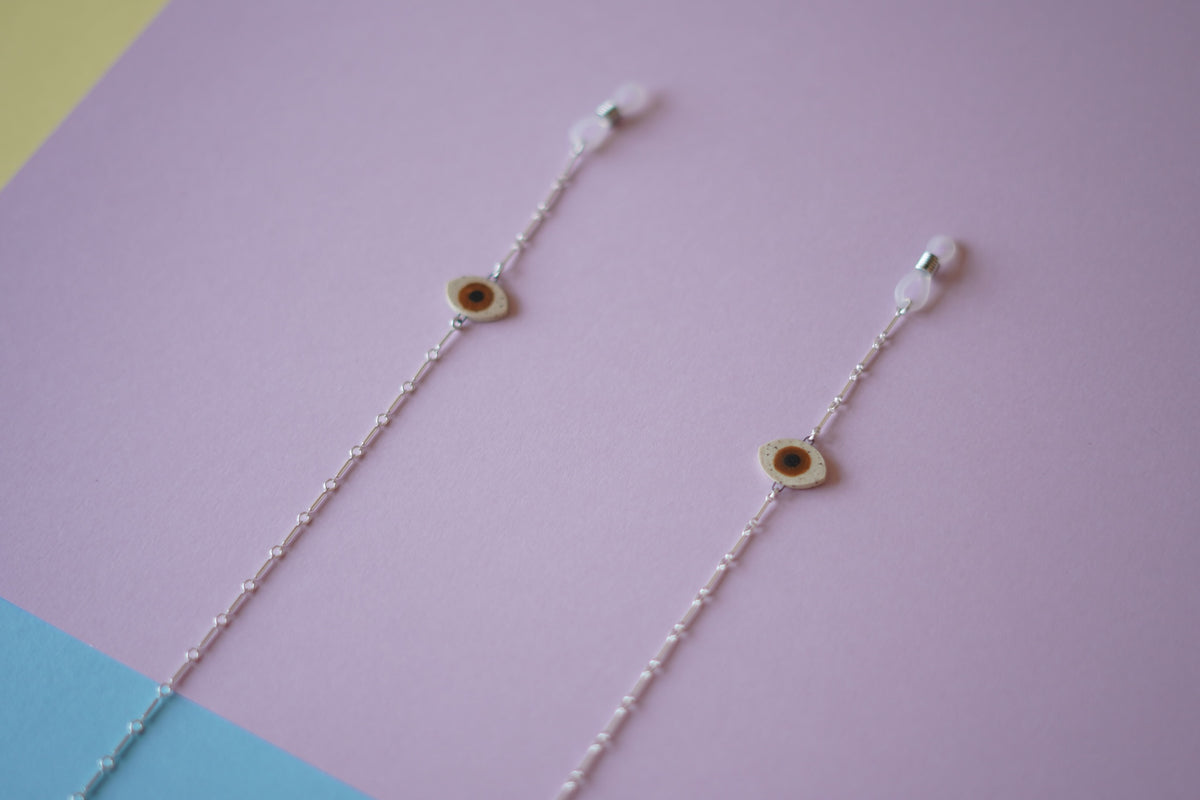 A pair of delicate silver chain earrings with small circular evil eye charms, displayed diagonally on a pastel purple background with blue and yellow corner accents.
