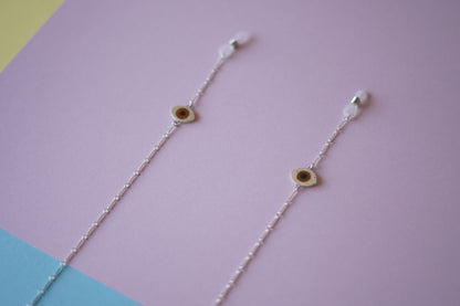 A pair of delicate silver chain earrings with small circular evil eye charms, displayed diagonally on a pastel purple background with blue and yellow corner accents.
